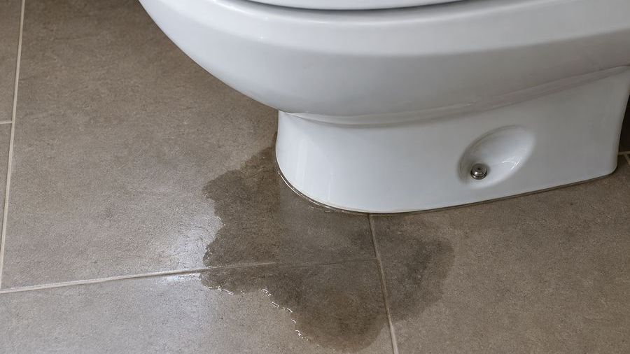 Poça de água se espalhando no piso ao redor da base de um vaso sanitário branco